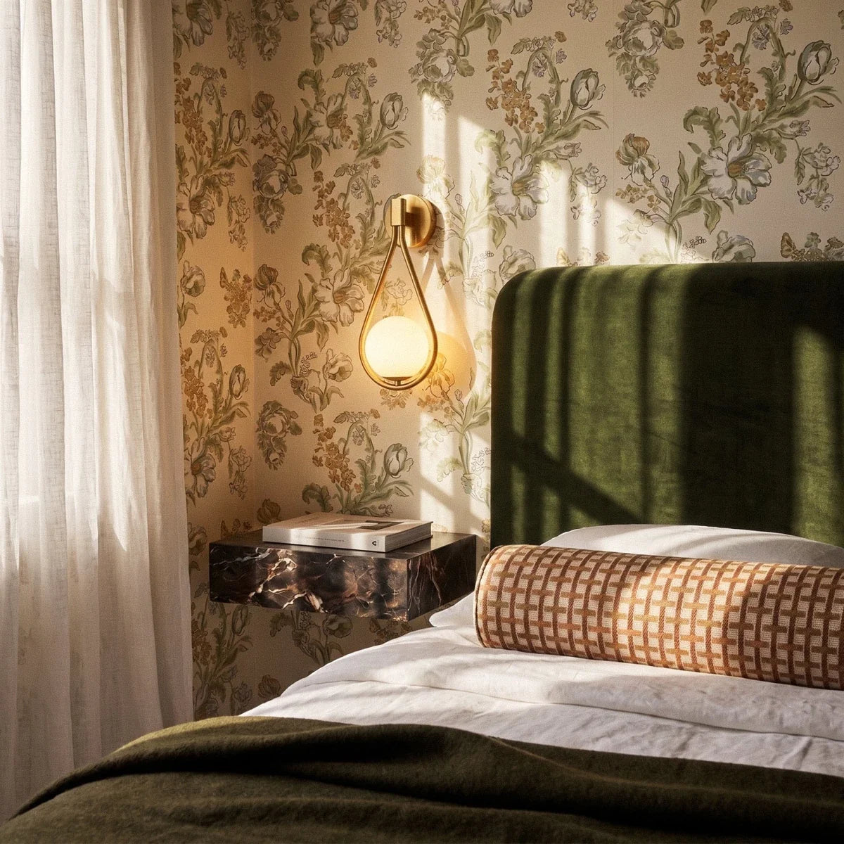 Clérachic gold iron arm wall sconce glowing beside a bed on floral wallpaper, above a dark marble nightstand.