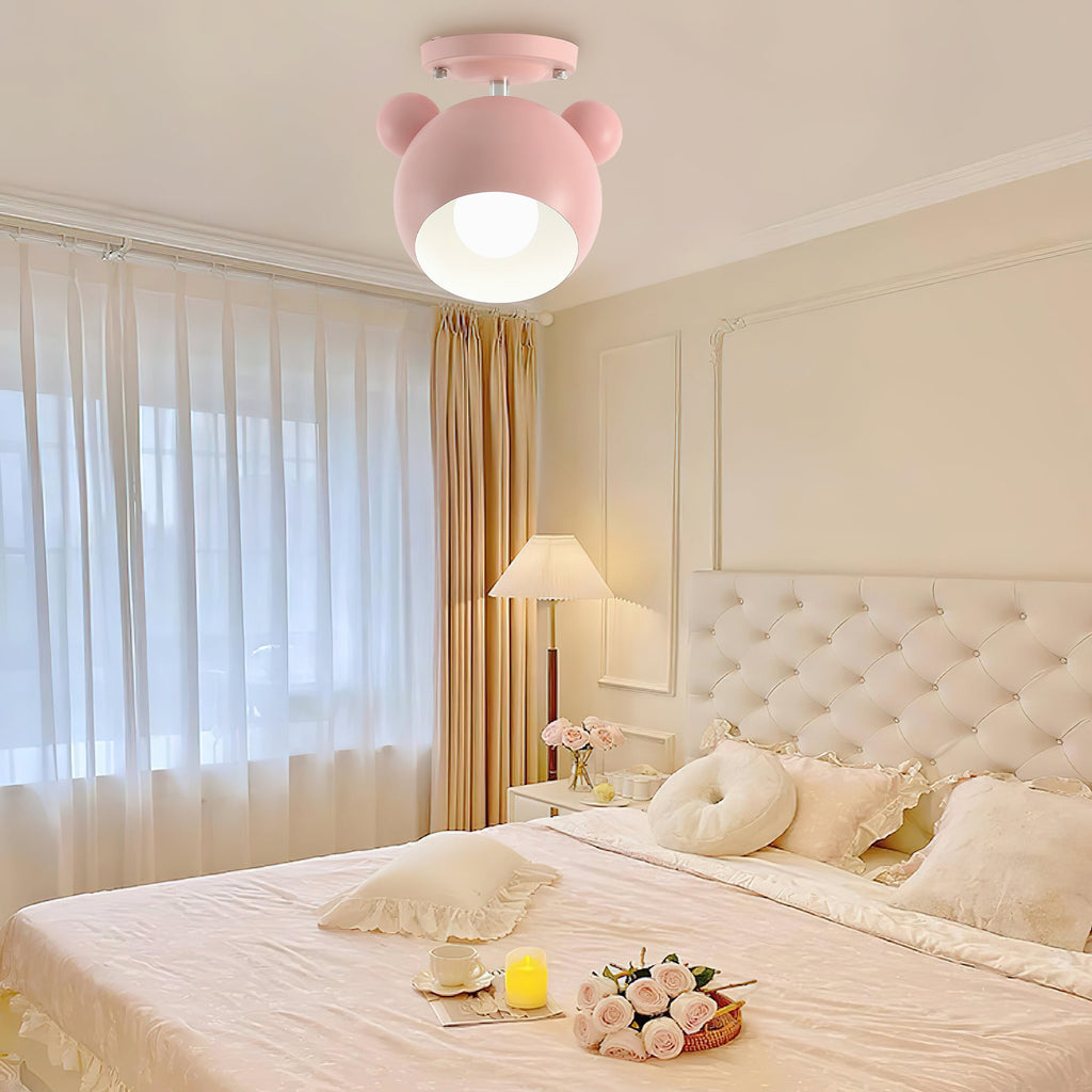 A pink Clérachic ceiling lamp lit above a cream bedroom with a tufted headboard, sheer curtains and a bedside lamp.