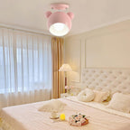 A pink Clérachic ceiling lamp lit above a cream bedroom with a tufted headboard, sheer curtains and a bedside lamp.