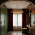 Clérachic brass ring chandelier glowing in a dressing room with green wardrobes, plum curtains and a boucle ottoman.
