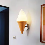 A lit Clérachic wall light shaped like a soft-serve cone, with a warm glow on a pale wall beside a dark doorway and a framed picture.
