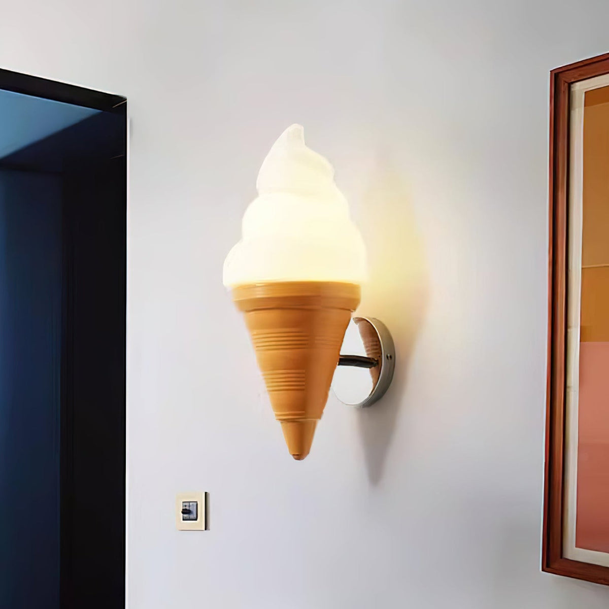 A lit Clérachic wall light shaped like a soft-serve cone, with a warm glow on a pale wall beside a dark doorway and a framed picture.