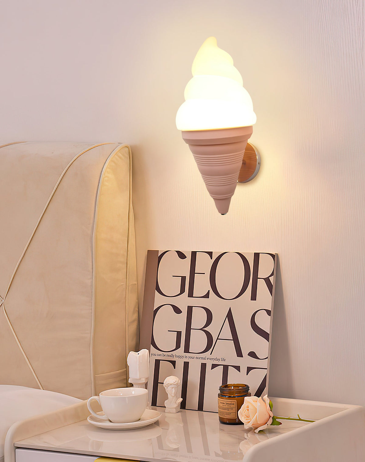 Pink-coned Clérachic wall light glowing above a bedside table with a teacup and an art book, next to a cream padded headboard.