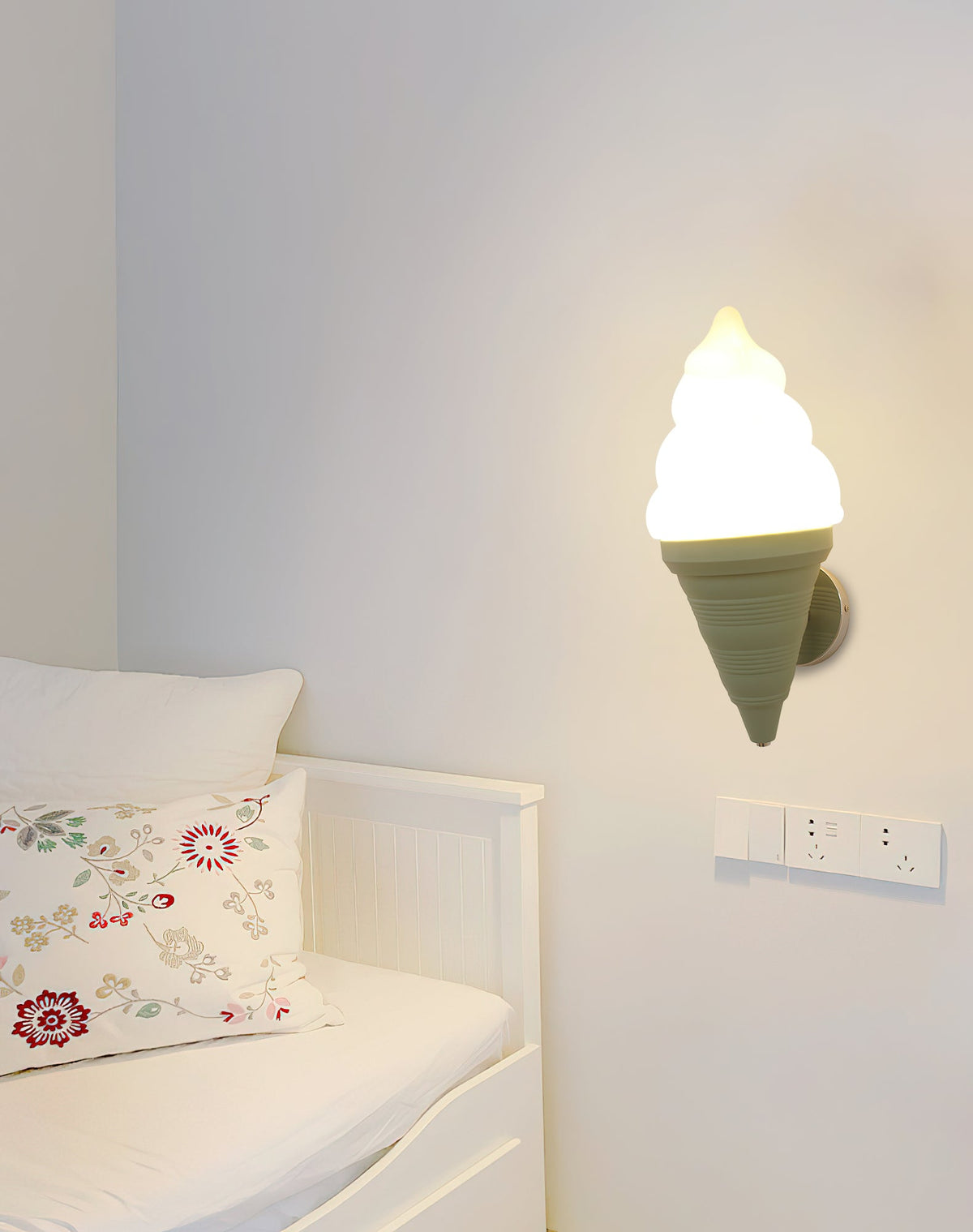 Green-coned Clérachic wall light lit beside a white daybed with a floral cushion and a wall socket plate on a pale wall.