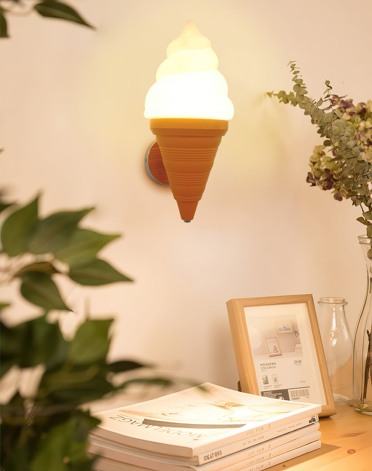 Orange-coned Clérachic wall light glowing over a desk with stacked magazines, a framed print and leafy plants.
