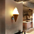Lit Clérachic wall light on a weathered concrete-look wall, with an open kitchen of dark wood cabinets behind it.