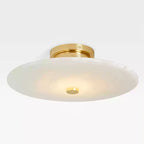 Clérachic flush mount seen at an angle, its alabaster disc lit warm beneath a polished brass ceiling collar.
