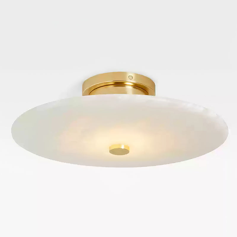 Clérachic flush mount seen at an angle, its alabaster disc lit warm beneath a polished brass ceiling collar.