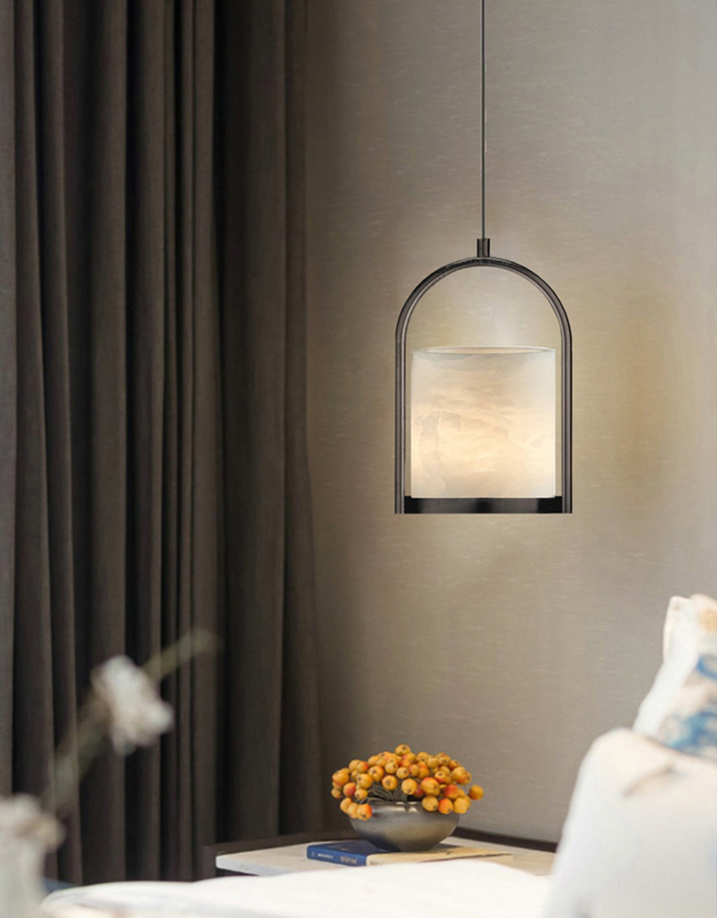The Clérachic Jewel Pendant Light lit beside heavy curtains, its pale alabaster shade framed by the dark arched stirrup.