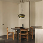 A Clérachic pendant in dark green glass hangs over a round dark table with rush-seat chairs, in a plain room with pale walls.