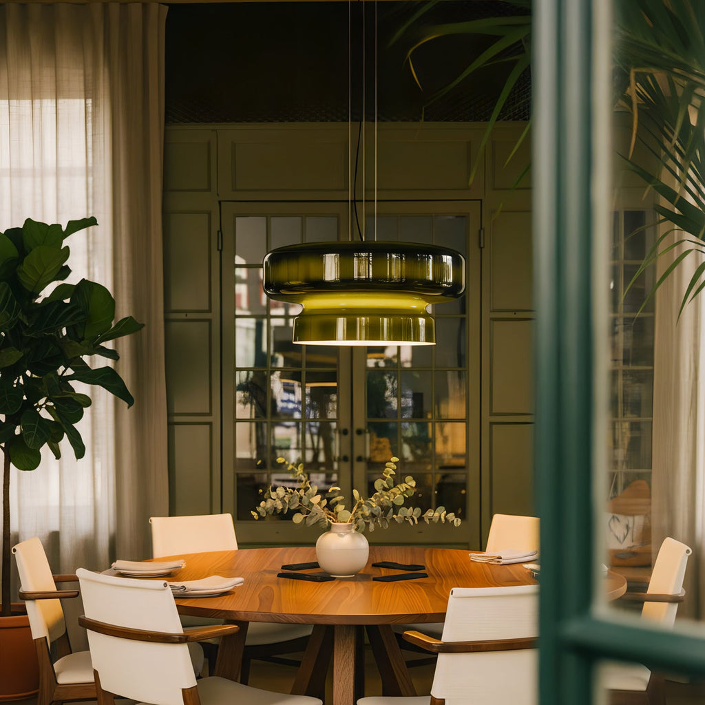 A lit Clérachic dark green glass pendant above a round wooden table with cream chairs, with green walls and a large leafy plant.