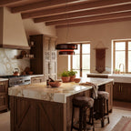 A Clérachic pendant in Amber Red glass above a marble-topped kitchen island with two dark stools, under timber ceiling beams.