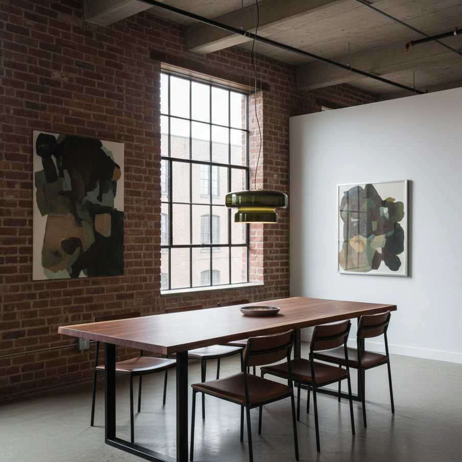 A Clérachic dark green pendant by a tall factory window, above a long dark wood table with slim chairs, against a brick and white wall.