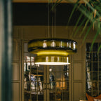 Close view of a Clérachic dark green glass pendant lit from below, with glass doors and a leafy plant behind it.