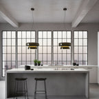 Two Clérachic dark glass pendants hang above a grey kitchen island with black stools, in front of a wall of grid-pane windows.
