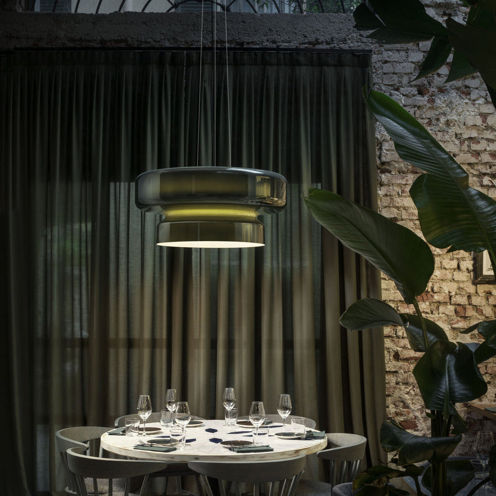 Clérachic dark green glass pendant behind sheer dark curtains, above a round white table set with glasses, with plants and a brick wall behind.