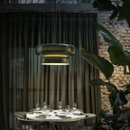 Clérachic dark green glass pendant behind sheer dark curtains, above a round white table set with glasses, with plants and a brick wall behind.