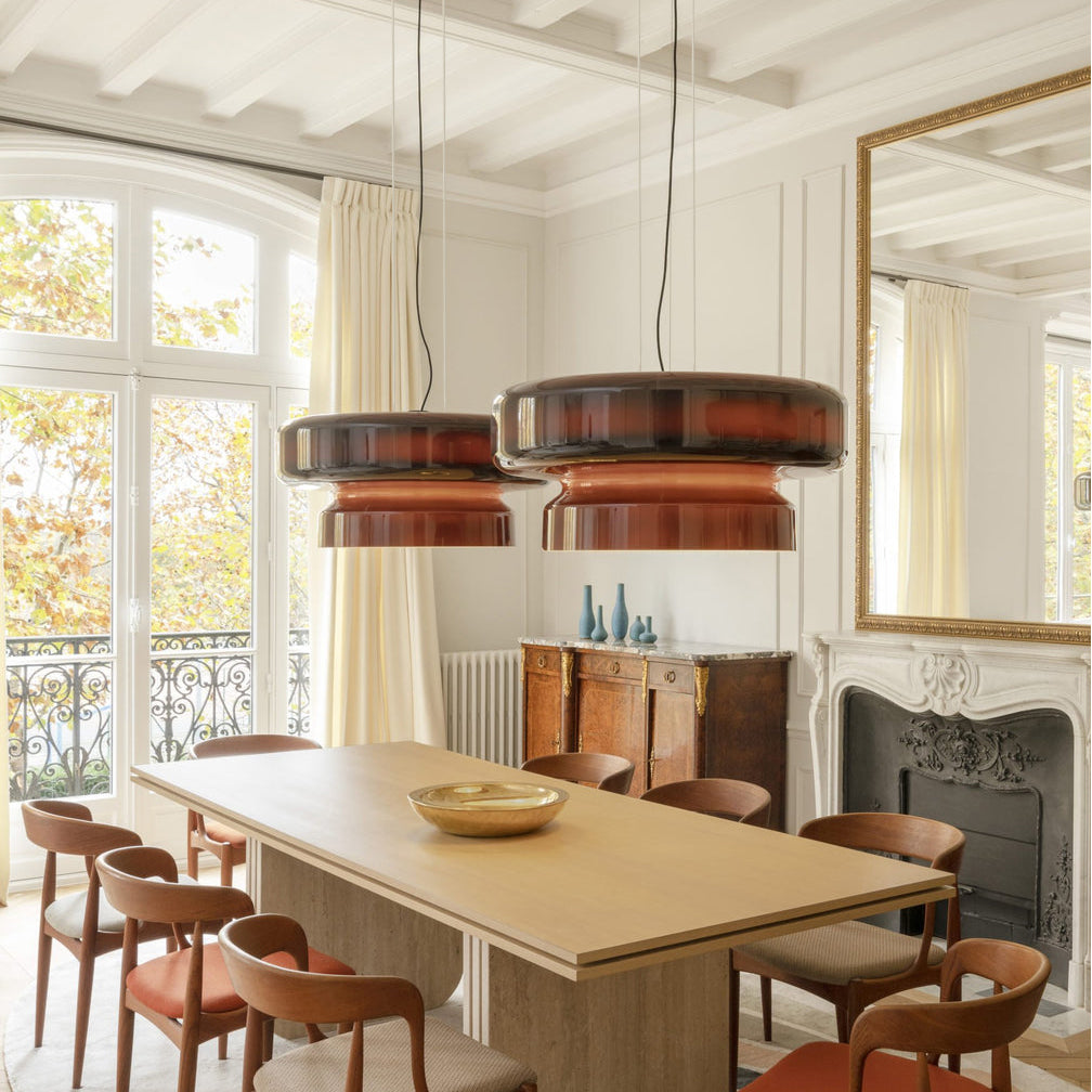 Close view of two Clérachic Amber Red glass pendants glowing in a bright dining room with tall windows and a gilt mirror.
