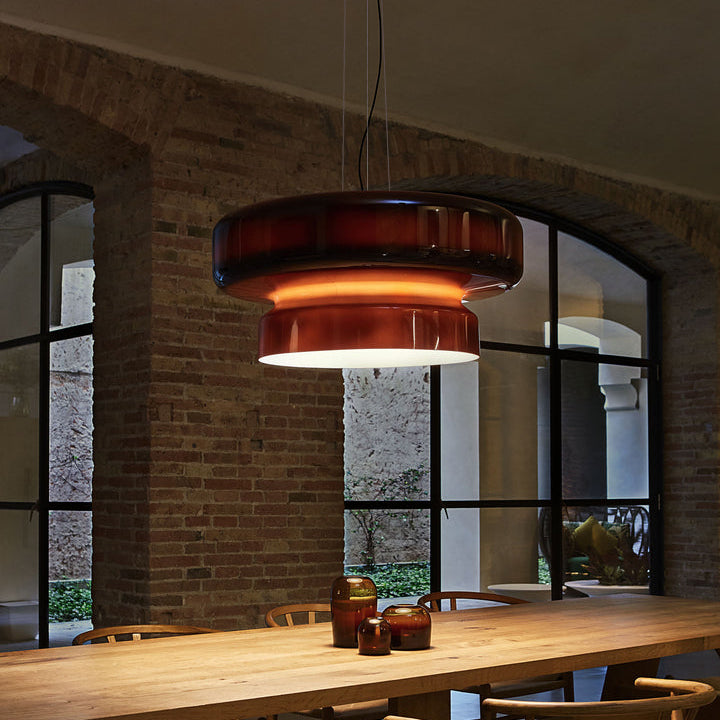 A Clérachic Amber Red glass pendant glows warm above a long timber table, in a brick room with arched black-framed windows.