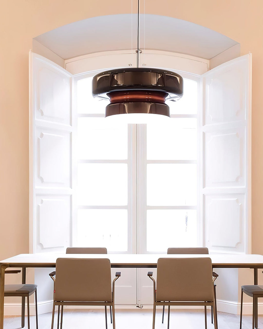 A Clérachic Amber Red glass pendant in front of white shutters and a window, above a table with grey-beige chairs.
