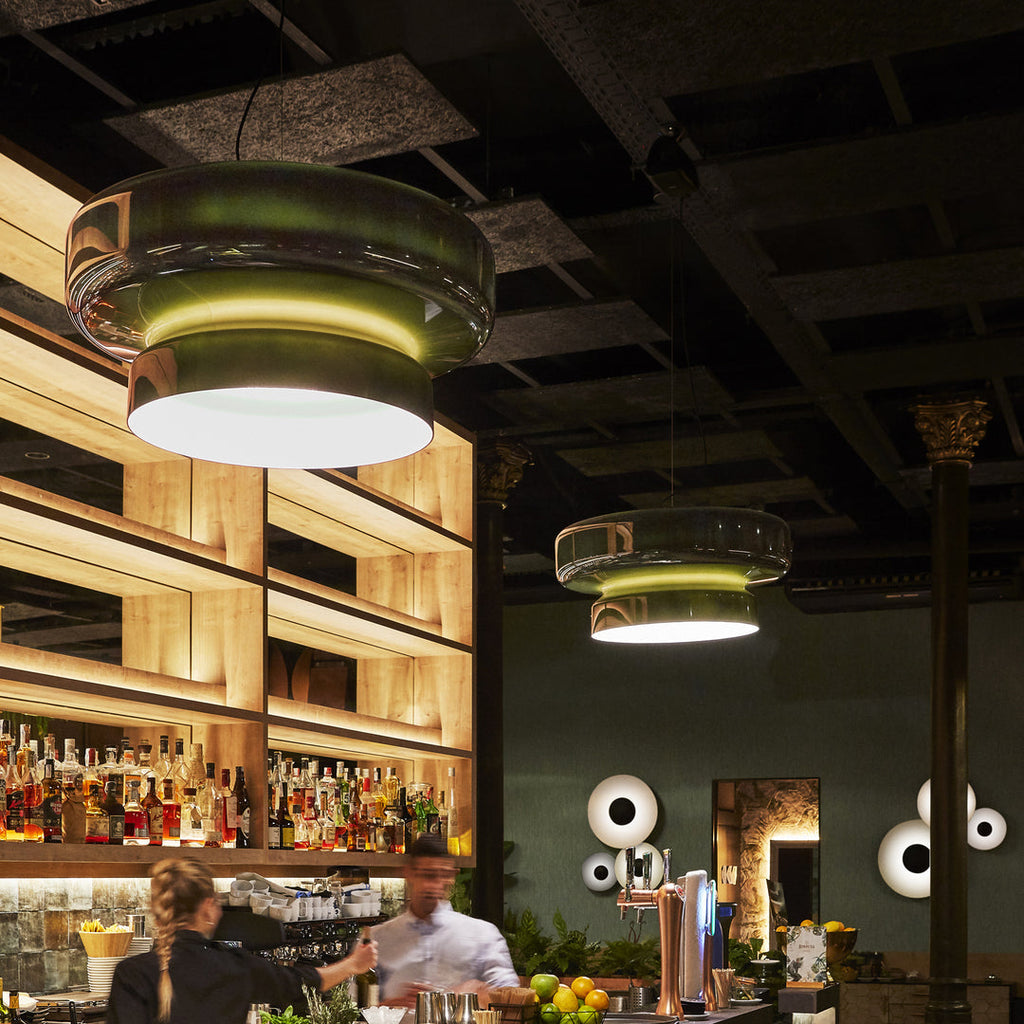 Clérachic dark green glass pendants glow in a restaurant bar, with lit shelves of bottles and bar staff in the background.