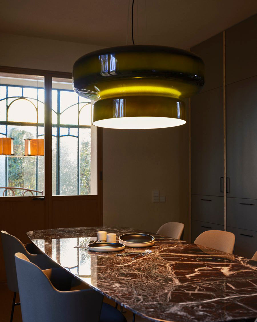 A Clérachic green glass pendant lit beside tall windows, above a marble dining table with blue chairs and a bowl on it.