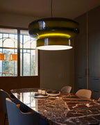 A Clérachic green glass pendant lit beside tall windows, above a marble dining table with blue chairs and a bowl on it.
