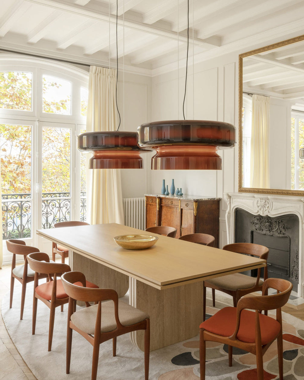 Two Clérachic Amber Red glass pendants above a pale dining table with russet chairs, with balcony windows and a fireplace behind.