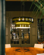 A Clérachic green glass pendant glows yellow-green above a wooden table, with arched glass doors and a plant behind.