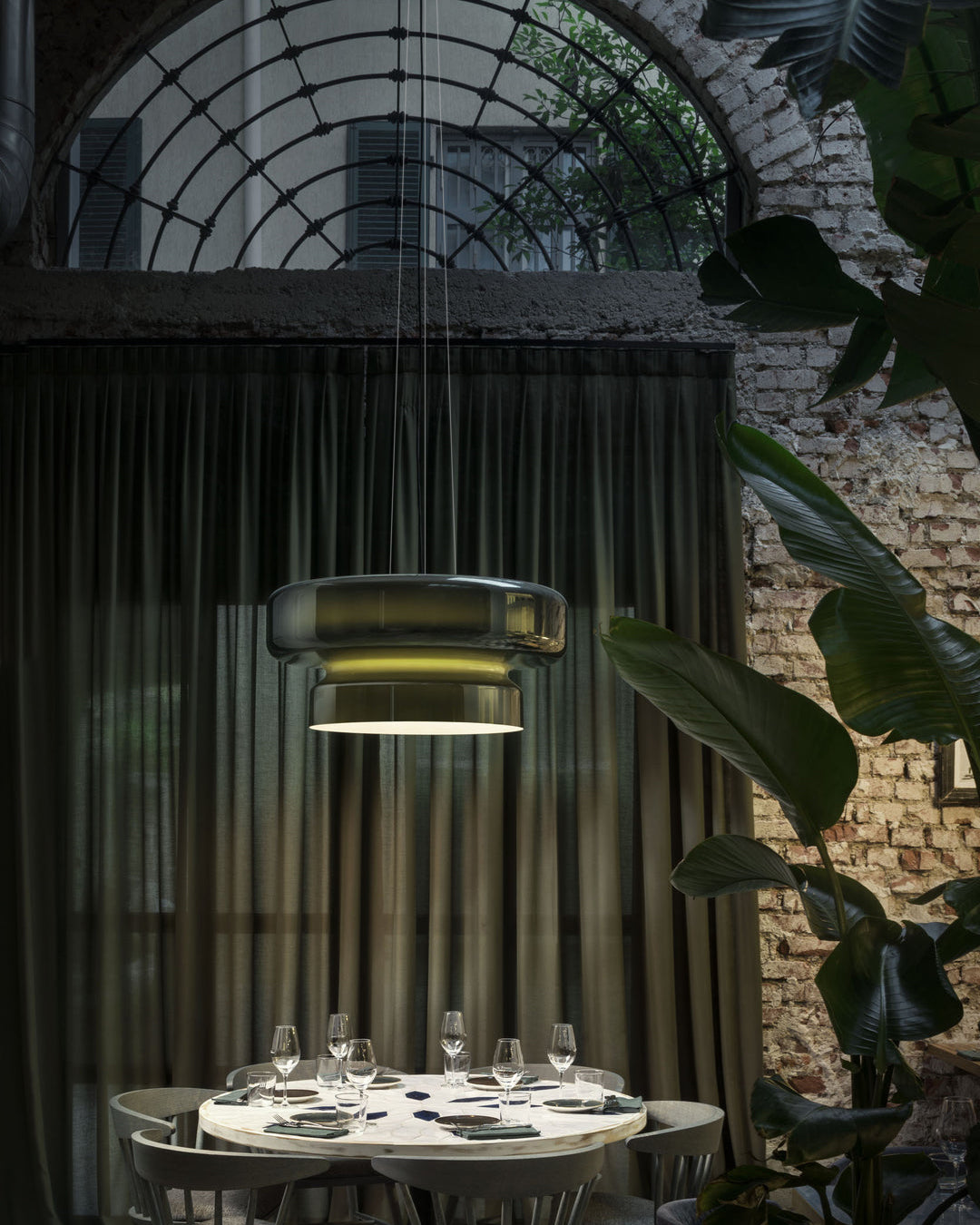 A Clérachic green glass pendant in a dark courtyard dining room, with sheer curtains, a brick wall and a table set for dinner.