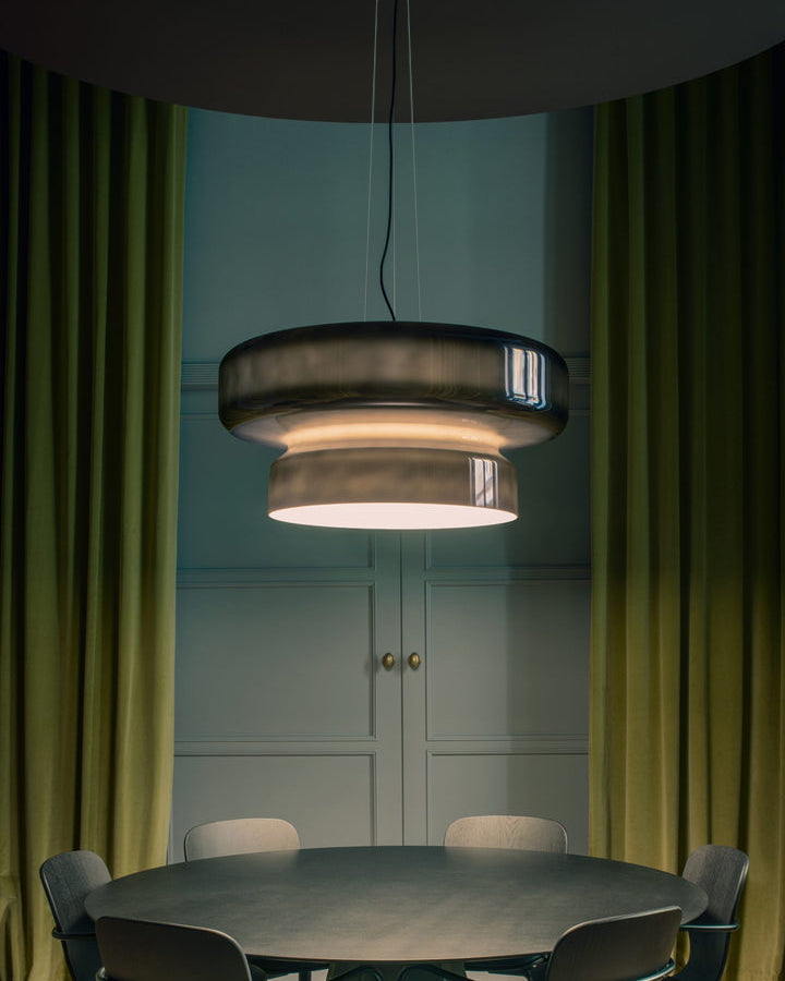 Clérachic smoky glass pendant between olive velvet curtains, above a round dark table in front of a panelled blue-green wall.