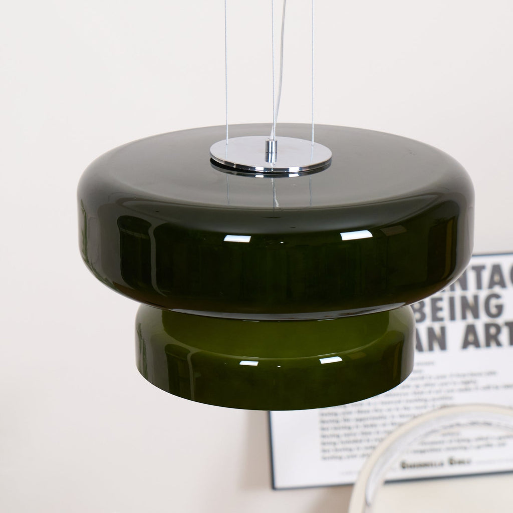 Close view of the Clérachic dark green pendant, with its chrome plate, wide glass disc and stepped lower tier, above a printed poster.