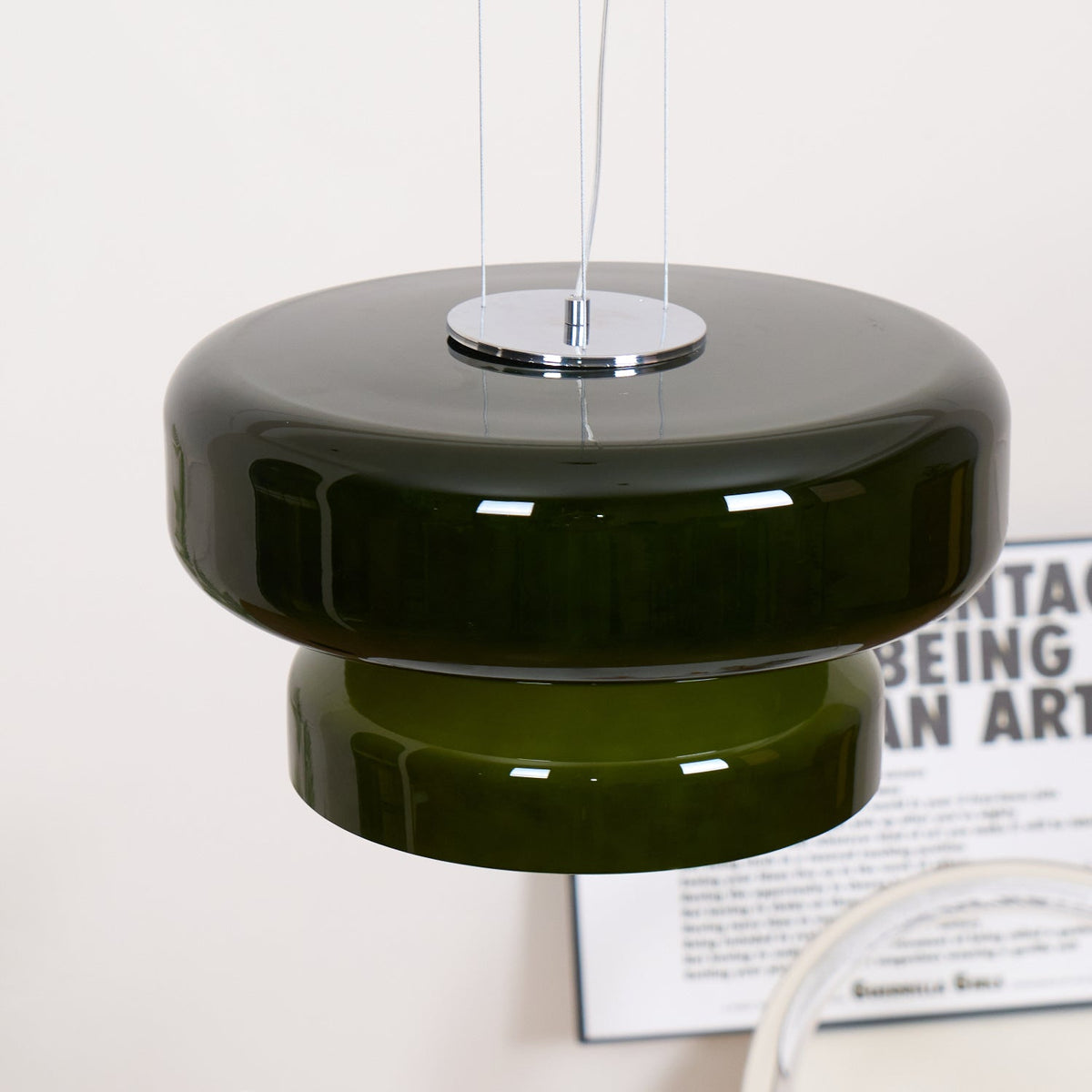 Close view of the Clérachic dark green pendant, with its chrome plate, wide glass disc and stepped lower tier, above a printed poster.
