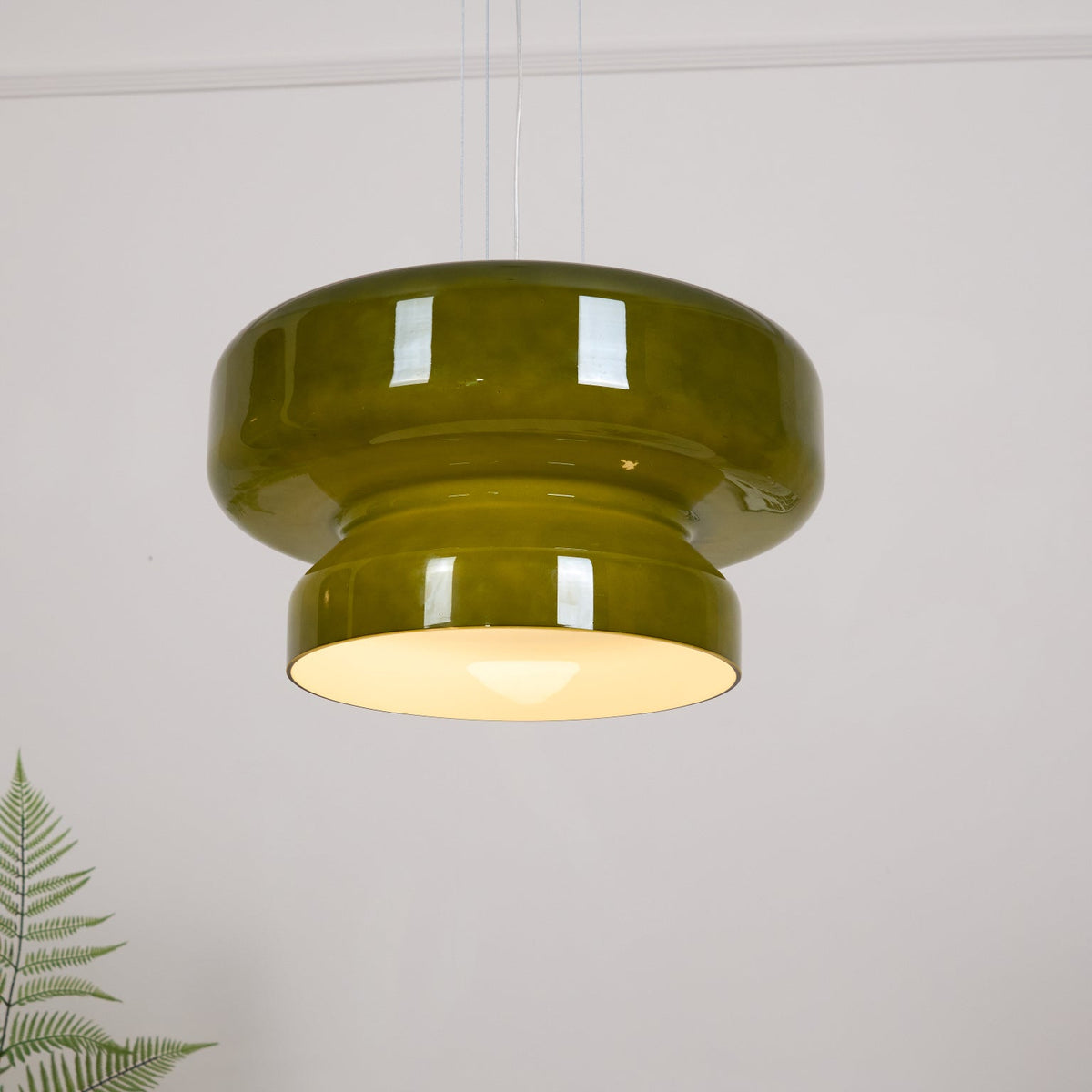 A lit Clérachic olive-green glass pendant beside a fern, its lower tier glowing warm white.