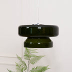 A Clérachic dark green glass pendant above a fern and a white table with a few small orange stems.
