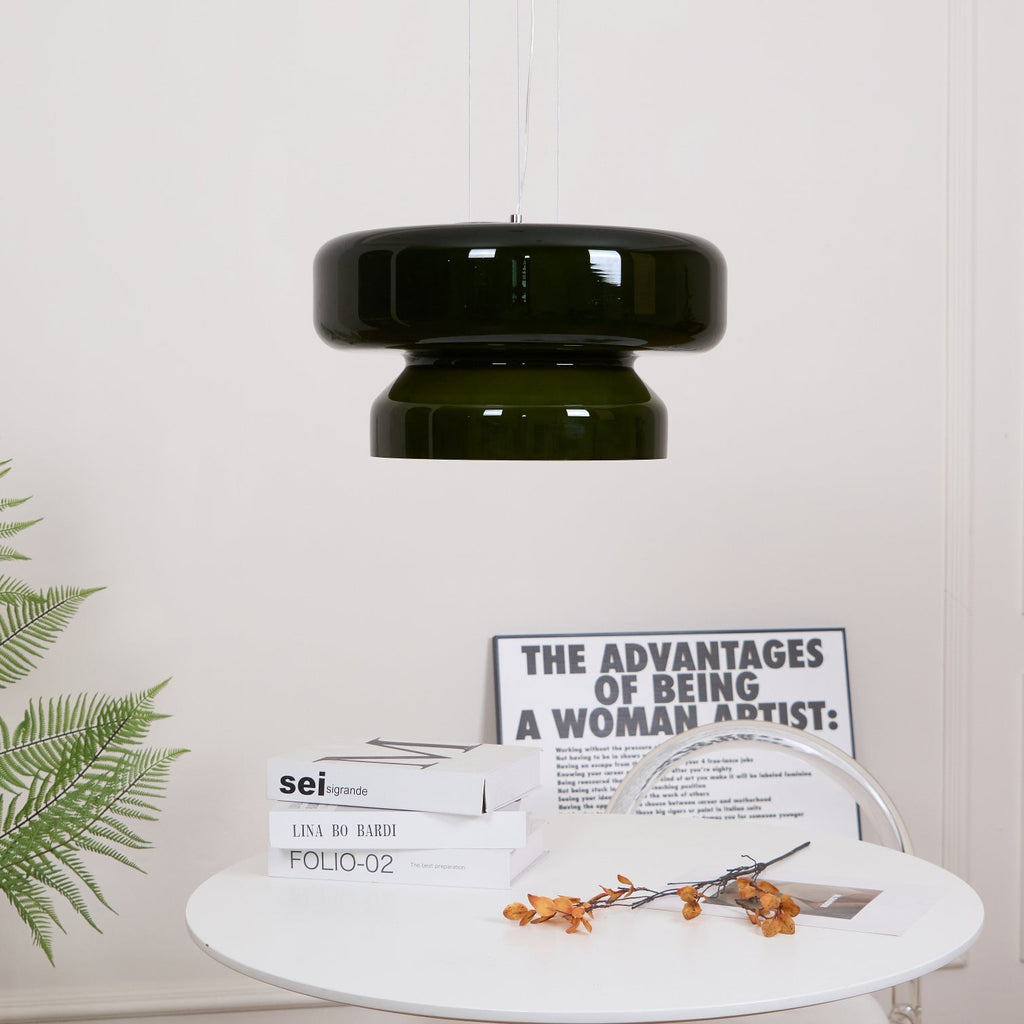 A Clérachic dark green glass pendant, small in the frame, above a round white table with books and orange stems.