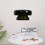 A Clérachic dark green glass pendant, small in the frame, above a round white table with books and orange stems.