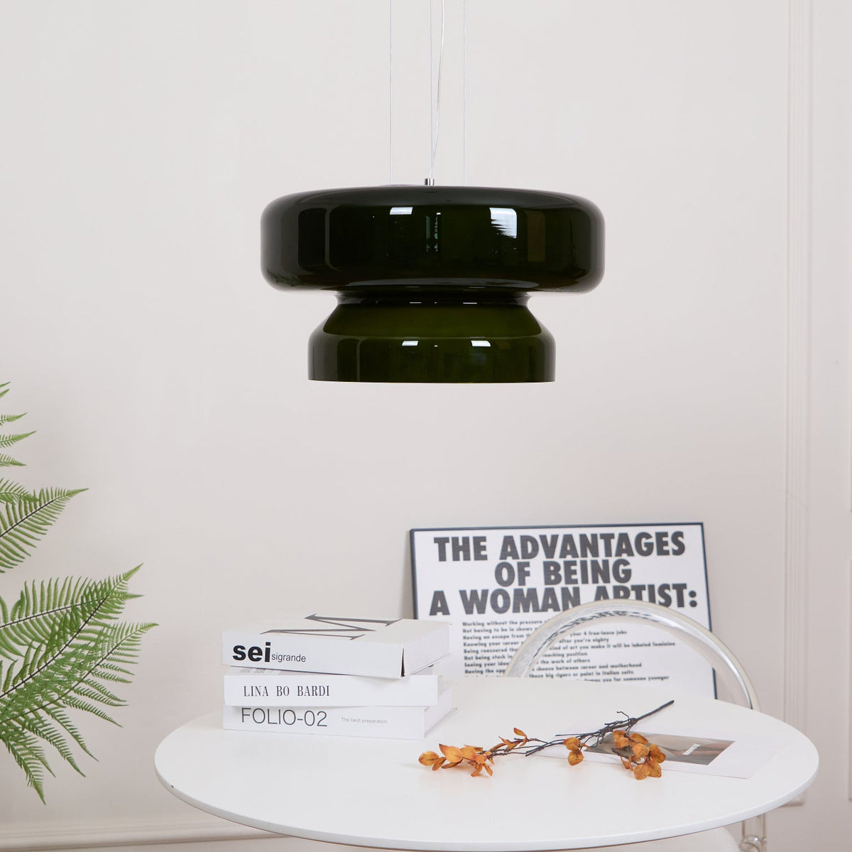 A Clérachic dark green glass pendant, small in the frame, above a round white table with books and orange stems.