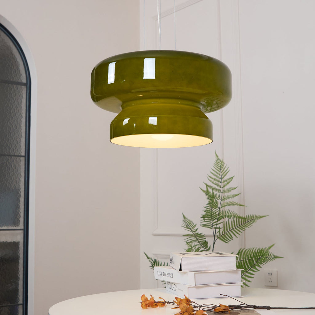 A lit Clérachic olive-green glass pendant beside a tall arched mirror, with a fern and a stack of books on a table.