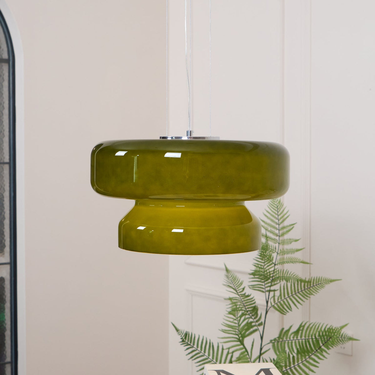 A Clérachic olive-green glass pendant with a chrome plate, above a fern and a white table.