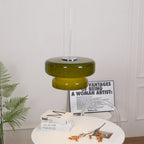 A Clérachic olive-green glass pendant, a little smaller in the frame, with books and a printed card on a table below.