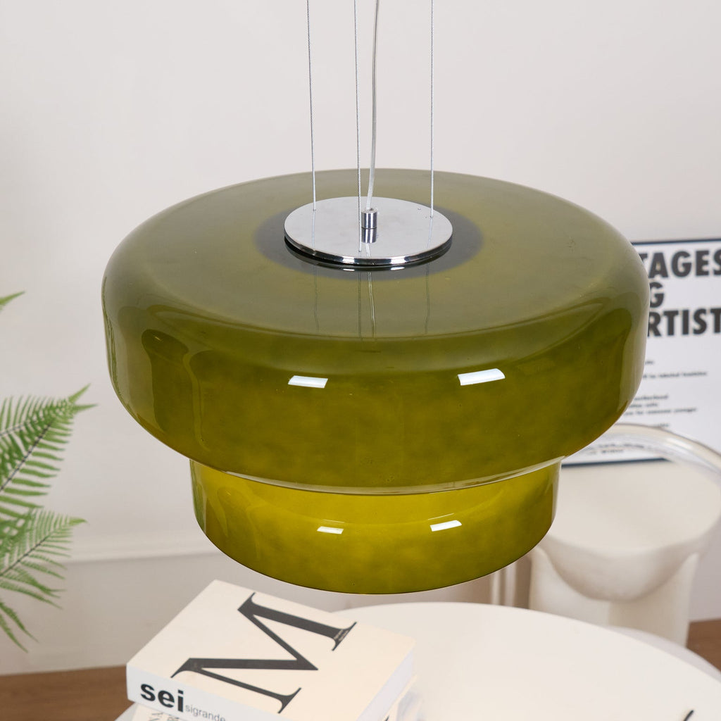 Close view of a Clérachic olive-green glass pendant, with its chrome top plate and fine cords, above a fern and a table.