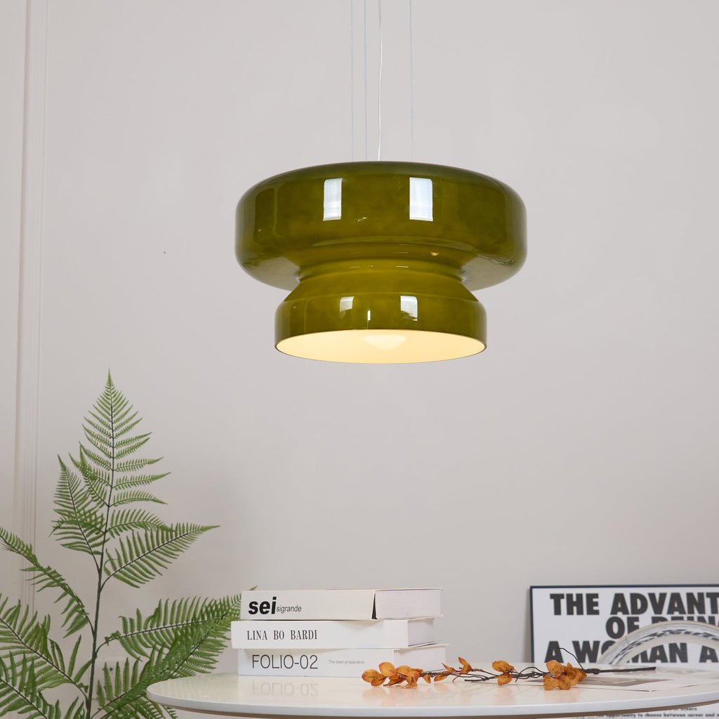 A lit Clérachic olive-green glass pendant above a fern and a round white table with books and orange stems.