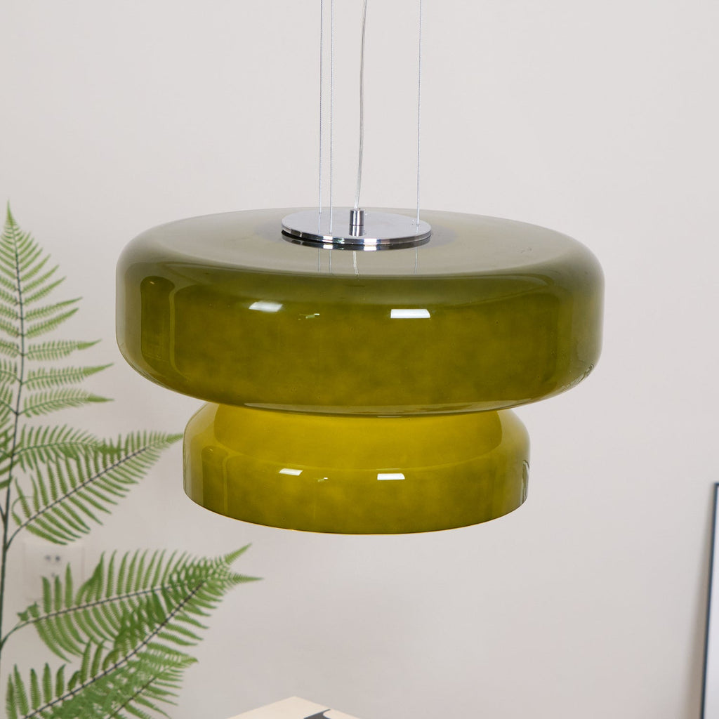 A Clérachic olive-green glass pendant with a chrome plate, in front of a white wall and above a fern.