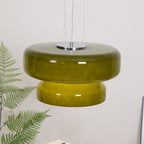 A Clérachic olive-green glass pendant with a chrome plate, in front of a white wall and above a fern.