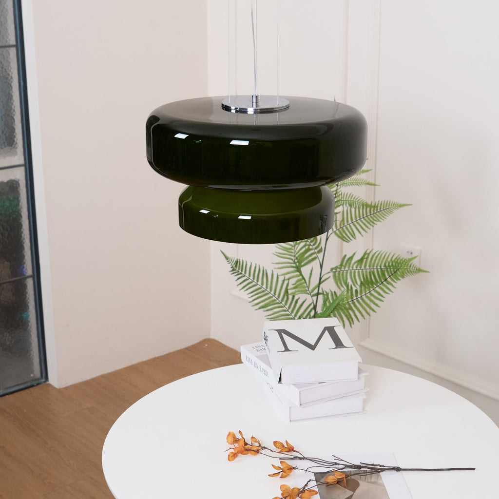 A Clérachic dark green glass pendant in a corner room with a wooden floor, above a fern and a round white table.