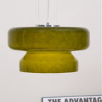 Close view of a Clérachic olive-green glass pendant against a white wall, with a poster edge at the bottom.