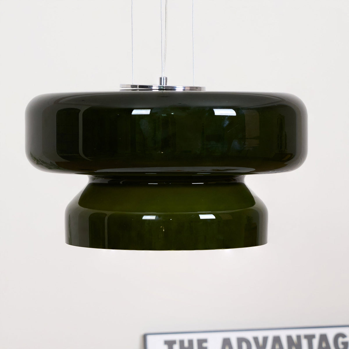 Close view of a Clérachic dark green glass pendant against a white wall, with its glossy upper disc and narrower tier.