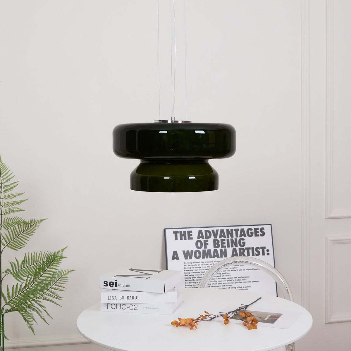 A Clérachic dark green glass pendant above a white table with books and orange stems, with a fern to the left.