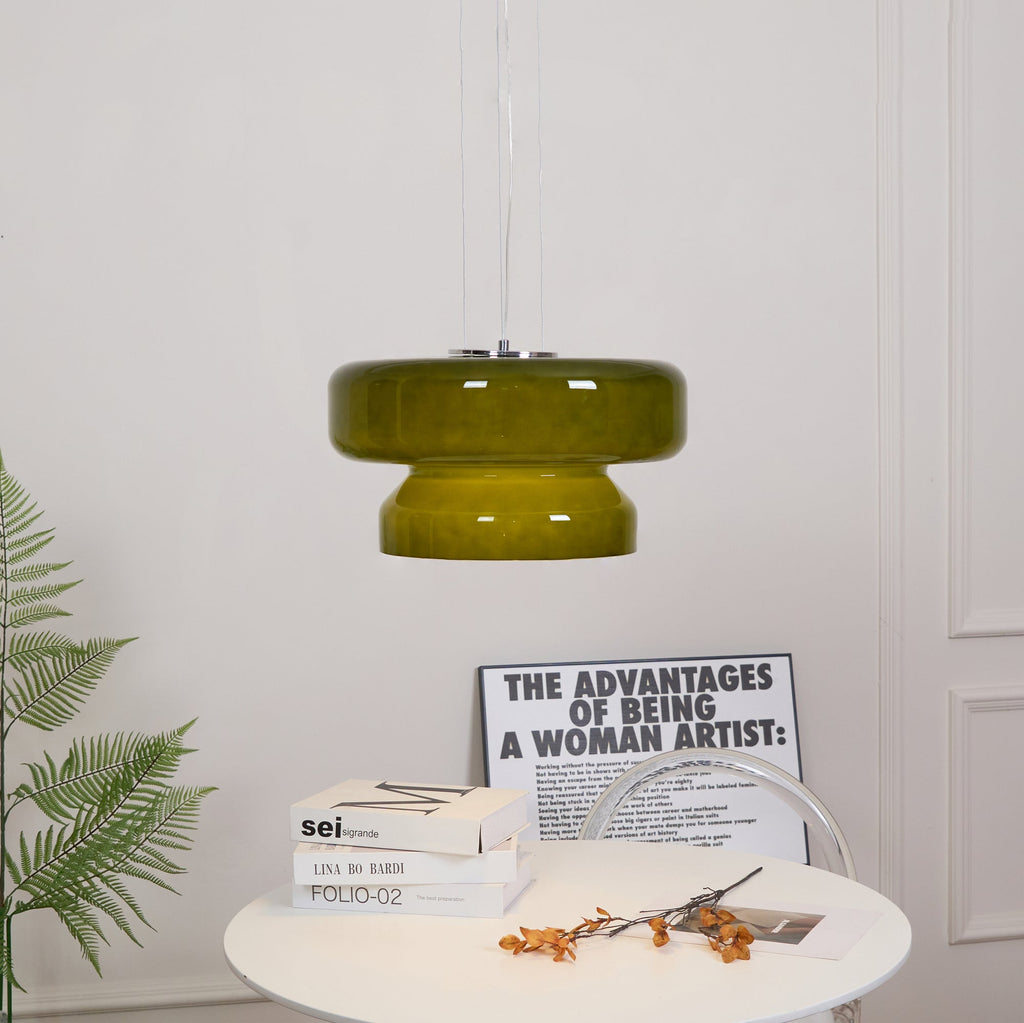 A Clérachic olive-green glass pendant above a white table with books, a printed card and orange stems, with a fern beside it.